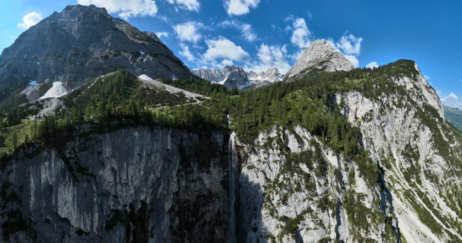 Ein Wasserfall stürzt von einer hohen Felswand, dahinter Bergpanorama | © TZA