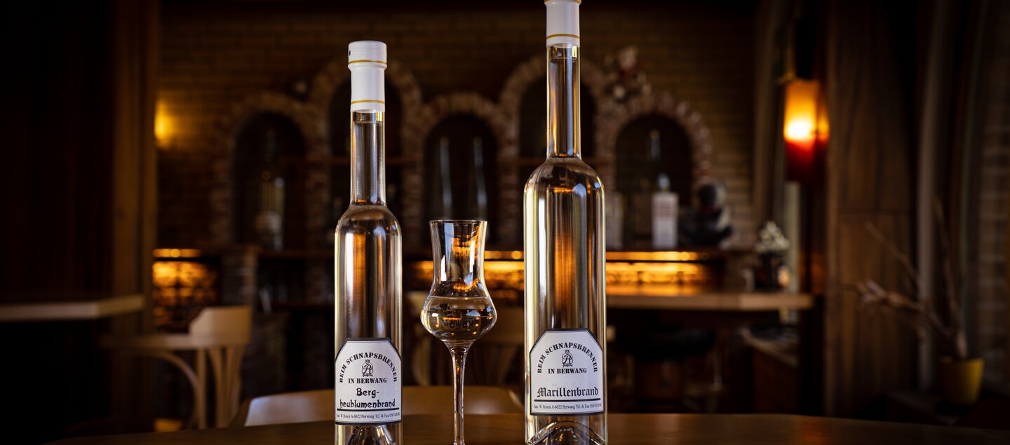 In the foreground, two bottles of schnapps and an elegant shot glass. A brick wall with dimmed light in the background. 