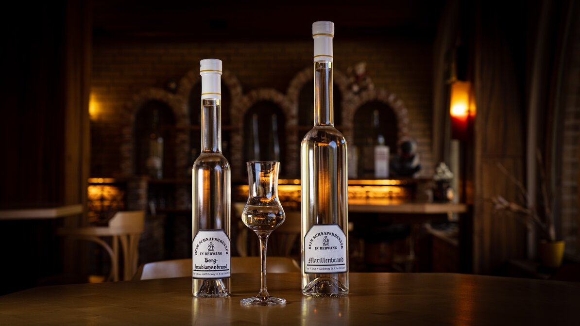 In the foreground, two bottles of schnapps and an elegant shot glass. A brick wall with dimmed light in the background. 