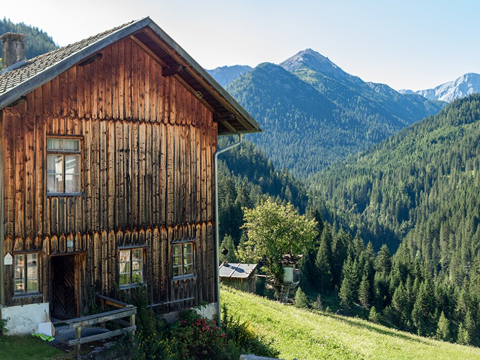 Ein altes Holzgebäude neben einer Wiese, im Hintergrund Bergpanorama | © Tiroler Zugspitz Arena/Bianca McCarty