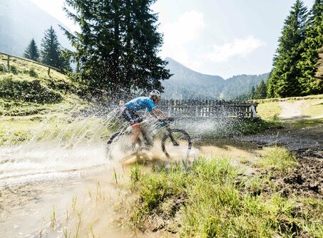 A mountain biker rides through a puddle, mountain panorama in the background | © Markus Greber