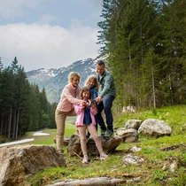 Eine Familie sieht sich etwas auf einem Smartphone an, sie stehen auf einer Lichtung im Wald, im Hintergrund Bergpanorama | © Tiroler Zugspitz Arena/Zotz Lea