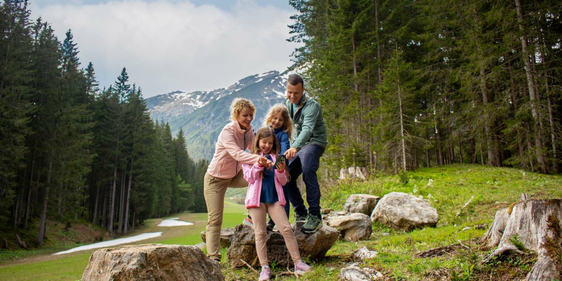 Eine Familie sieht sich etwas auf einem Smartphone an, sie stehen auf einer Lichtung im Wald, im Hintergrund Bergpanorama | © Tiroler Zugspitz Arena/Zotz Lea