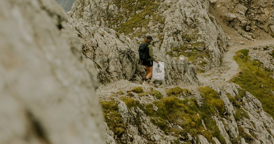 A young woman is walking along a hiking trail in the rocks. She is holding on to a steel cable on the left. In her right hand she is holding a bag for collecting garbage.  | © Patron Plasticfree Peaks _ Lena Everding Photo