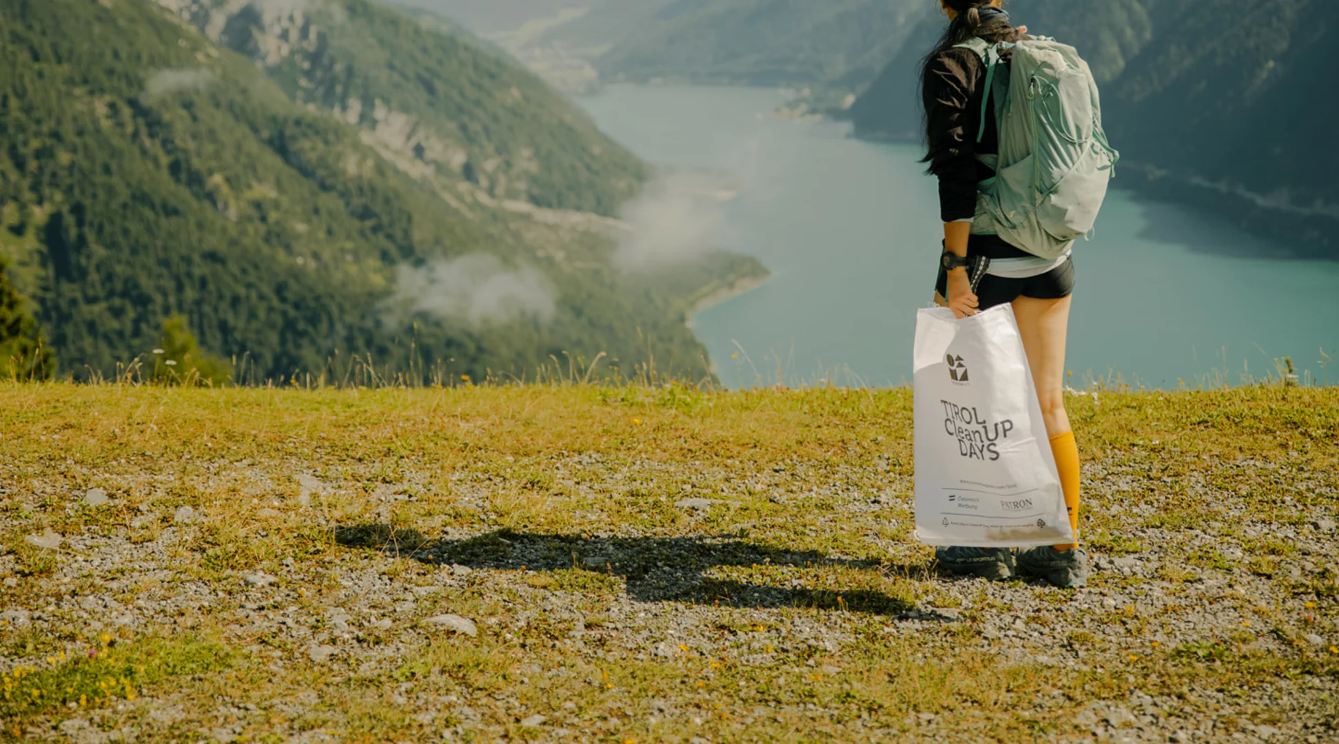 Clean Up Days Tirol | © Patron Plasticfree Peaks _ Lena Everding Photo Woman looking out over a beautiful lake from a grassy hill, holding the Clean Up Days garbage collection bag. | © Patron Plasticfree Peaks _ Lena Everding Photo