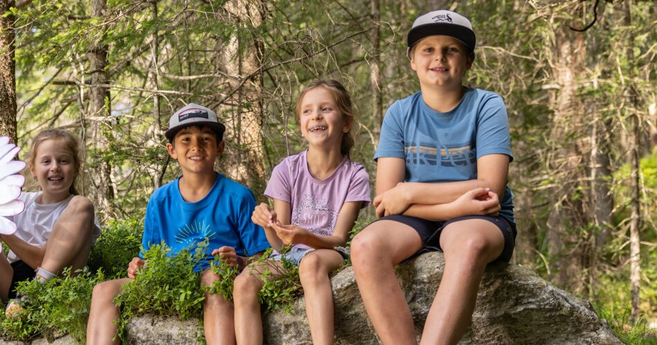 Vier Kinder sitzen lachend auf einem Stein in einem schönen Wald, ein Mädchen hält ein großes, lachendes, künstliches Gänseblümchen in der Hand | © Johannes Sautner / Shootandstyle