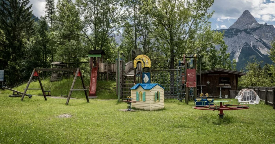 Spielplatz auf einer Wiese vor Bäumen und Bergpanorama | © TZA