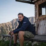 Man on a terrace in front of a wooden house. Mountains in the background. | © Heinzlmeier