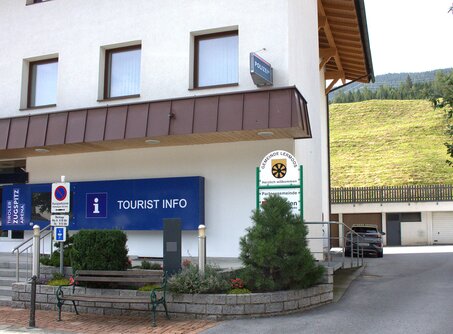 A house with the Tiroler Zugspitz Arena logo and the Tourist Info sign next to it. A driveway can be seen on the right, where a gray car is parked. | © TZA