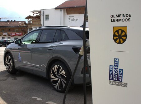 A gray car parked in front of a white electric charging station with the logo of the Tiroler Zugspitz Arena and the logo of the municipality of Lermoos | © TZA