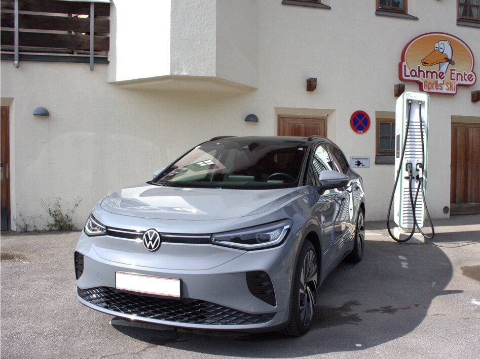 A gray car parked by a white electric charging station in front of a house with the sign Lahme Ente Apres Ski  | © TZA