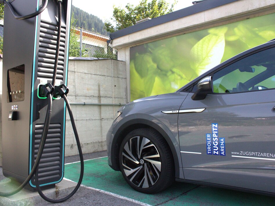 A gray car parked next to a black e-charging station, a green poster with a leaf in the background | © TZA