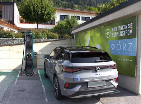 A gray car is parked next to a black e-charging station, in the background a green poster with a leaf and the inscription, here you fill up with solar power, Wörz. | © TZA