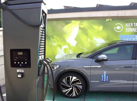 A gray car parked next to a black e-charging station, a green poster with a leaf in the background | © TZA