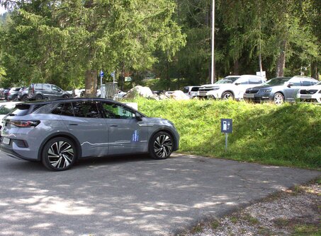 A gray car, next to it a sign with the symbol of an e-charging station. A small patch of grass in front of the car. | © TZA
