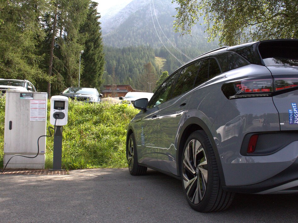 Ein graues Auto, daneben eine weiße E-Ladesäule. Im Hintergrund ist ein Abschnitt der Tiroler Zugspitzbahn zu sehen. | © TZA