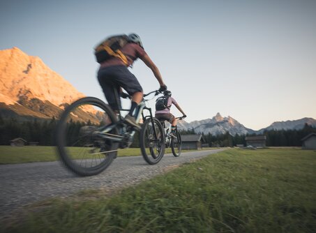 Zwei Radfahrer auf einem Weg, dahinter ein von der Abendsonne angestrahlter Berg | © Tiroler Zugspitz Arena/C. Jorda