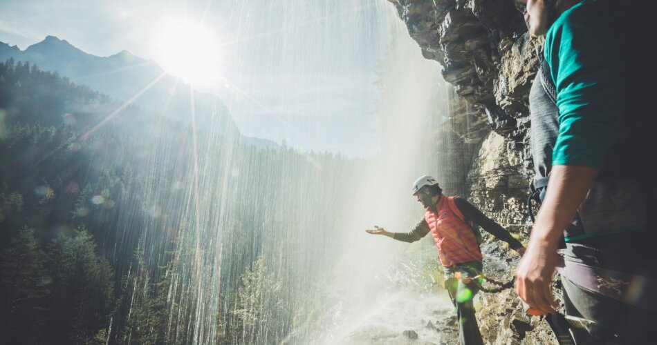 Ein Mann und eine Frau gehen gesichert am Seil unter einem Wasserfall hindurch. | © Tiroler Zugspitz Arena/C. Jorda