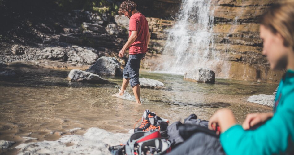 Two people, a man walks barefoot through a small pool of water at the foot of a waterfall, a woman sits on the edge | © Tiroler Zugspitz Arena/C. Jorda