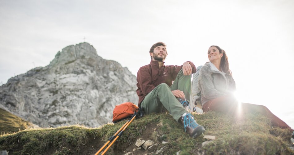 Two hikers sitting on an alpine meadow, a rocky peak in the background | © Tiroler Zugspitz Arena/C. Jorda