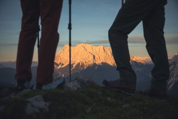 Im Vordergrund die Beine von zwei Wanderern, im Hintergrund die Zugspitze im Abendlicht | © Tiroler Zugspitz Arena/ C. Jords