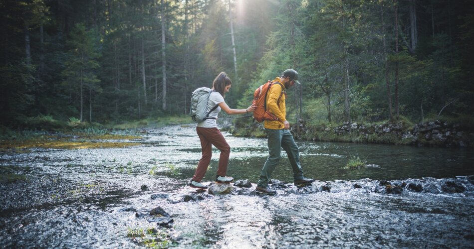 Zwei Wanderer spazieren auf einer Waldlichtung, an einer sehr flachen Stelle, durch einen schönen Fluss. | © Tiroler Zugspitz Arena/ C.Jorda