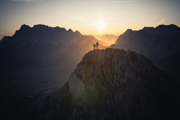 Zwei Personen wandern in der Morgensonne, im Hintergrund Bergpanorama und die aufgehende Sonne | © TZA/ C. Jorda