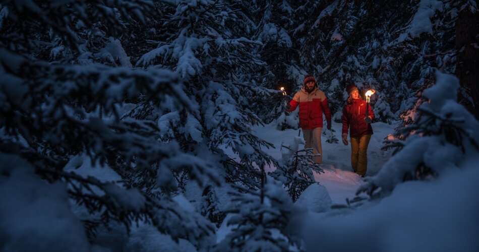 Ein Mann und eine Frau zur Dämmerung mit Fackeln im verschneiten Tannenwald. | © TZA_C. Jorda