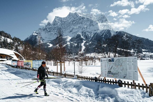 A cross-country skier, a trail sign with a map, and the Zugspitze | © Tiroler Zugspitz Arena/ Roast Media