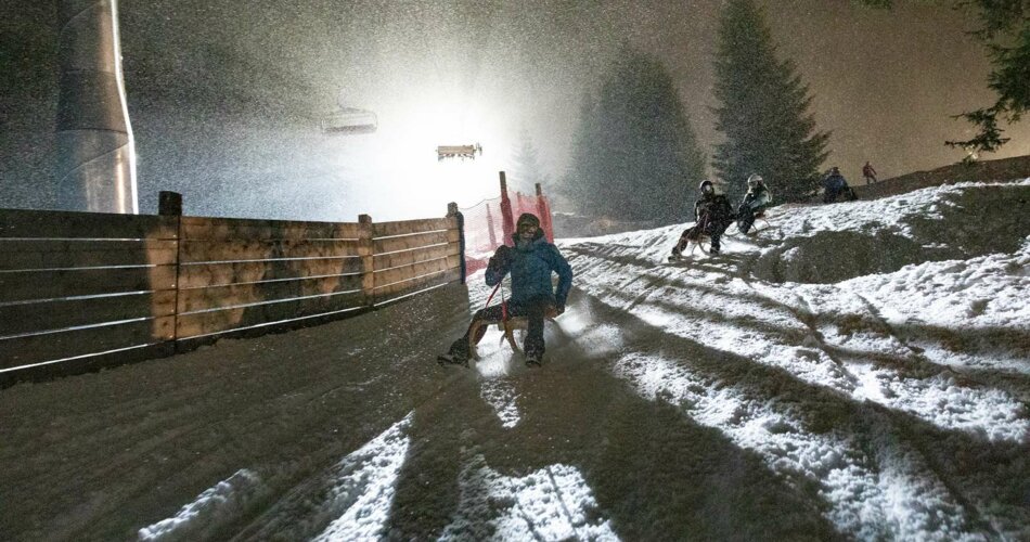 Eine Familie beim Nachtrodeln in Berwang, die Rodelbahn ist beleuchtet, es schneit | © Tiroler Zugspitz Arena/ Valentin Schennach