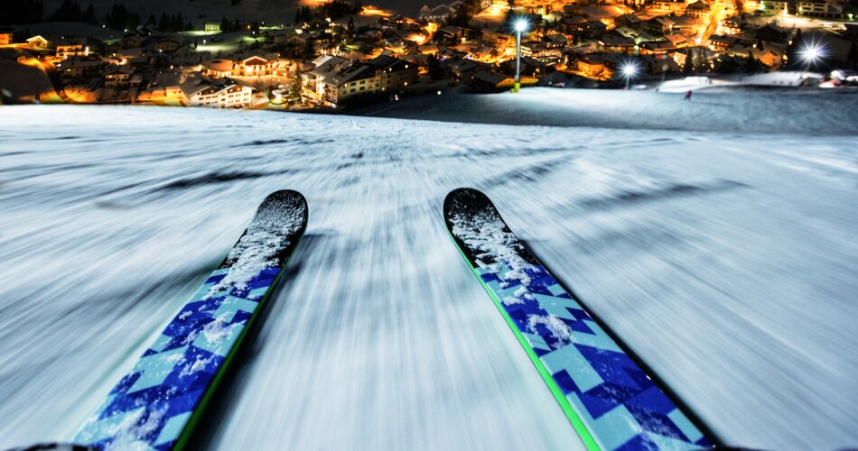 Ein während des Fahrens aufgenommenes Foto von einem Paar Ski, im Hintergrund ein beleuchtetes Dorf in der Nacht | © Tiroler Zugspitz Arena, C.Jorda