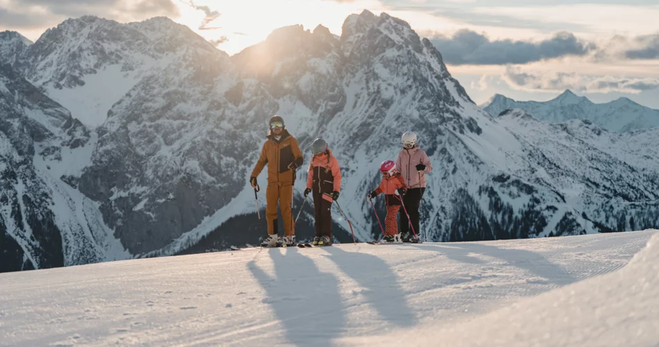 Dreiköpfige Familie bei blauem Himmel zur golden hour auf der Piste am Grubigstein. Die Berge Schartenkopf und Drachenkopf im Hintergrund.  | © TZA / Sam Oetiker
