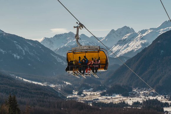 A family sits on a chairlift and waves, with a mountain panorama behind them | © Tiroler Zugspitz Arena/Sam Oetiker