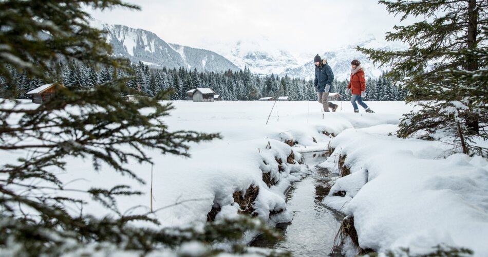 Two people hike through deep snow, a small stream beside them, a snowy winter landscape in the background | © TZA/ C.Jorda