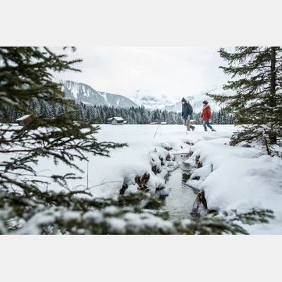 Zwei Personen stapfen durch tiefen Schnee, daneben ein kleiner Bach, im Hintergrund eine verschneite Winterlandschaft | © TZA/ C.Jorda