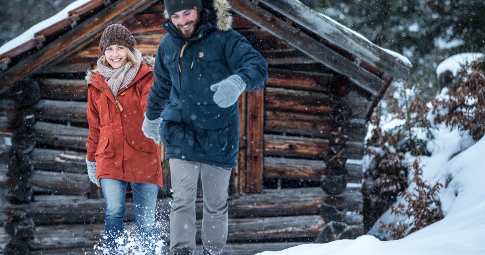 A couple holding hands and laughing as they walk through the deep snow, a wooden hut in the background | © TZA/ C.Jorda