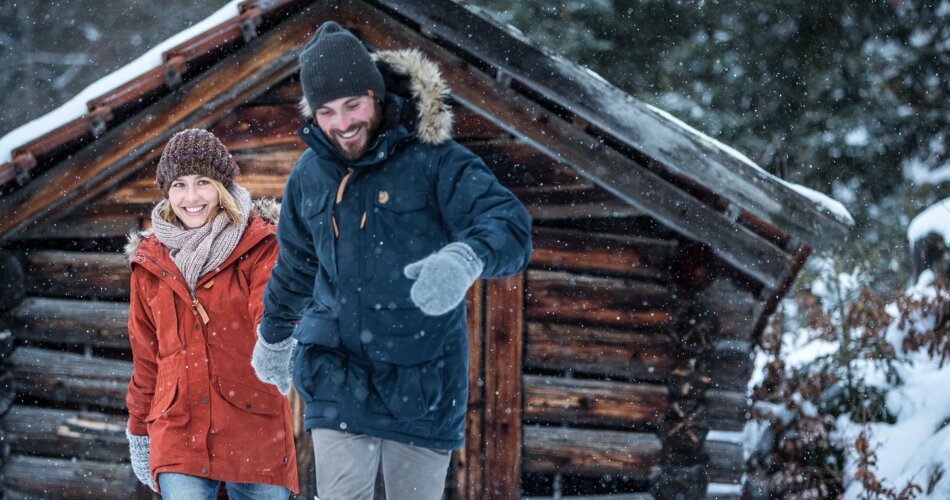 Ein Pärchen hält sich an den Händen und läuft lachend durch den tiefen Schnee, im Hintergrund eine Holzhütte | © TZA/ C.Jorda