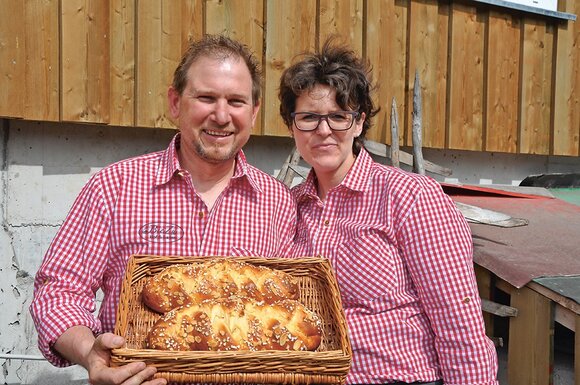 A man and a woman in red and white checked shirts, holding a woven basket with two yeast plaits in it. 