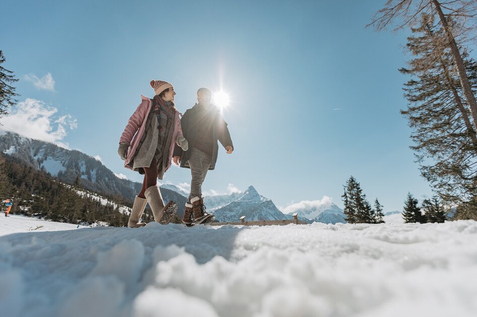 Snow-covered hill on which a man and a woman are walking, in the background the snow-covered Zugspitze, slightly shrouded in clouds. | © (c)ZABT_C. Jorda (66)