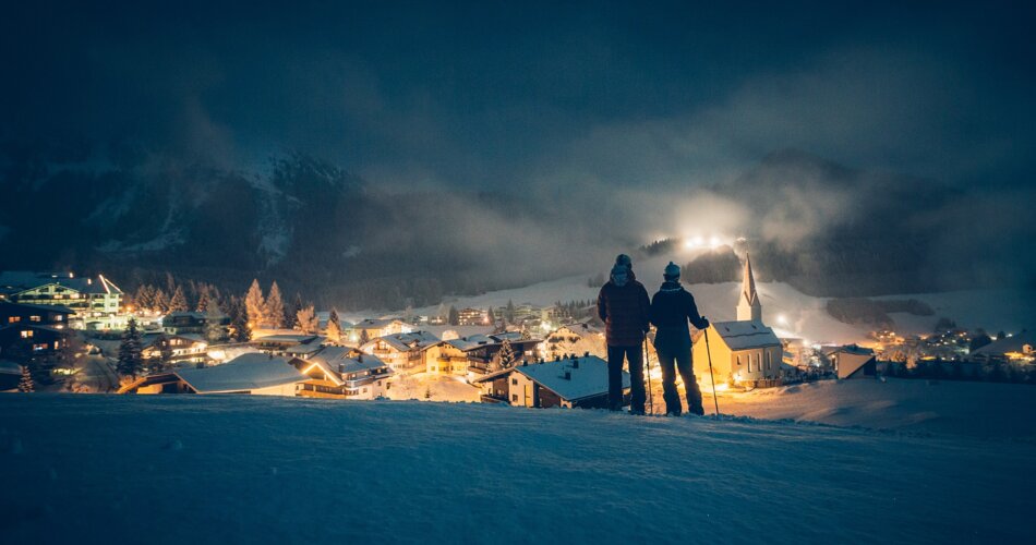 Zwei Personen machen eine Winterwanderung, es ist bereits dunkel, im Hintergrund sind die Lichter eines verschneiten Bergdorfes zu sehen | © Tiroler Zugspitz Arena