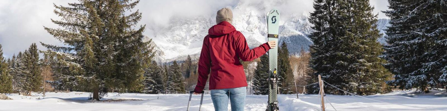 Young woman with a suitcase and skis next to her walks towards the Zugspitze which is visible in the background. The landscape is covered in snow. 