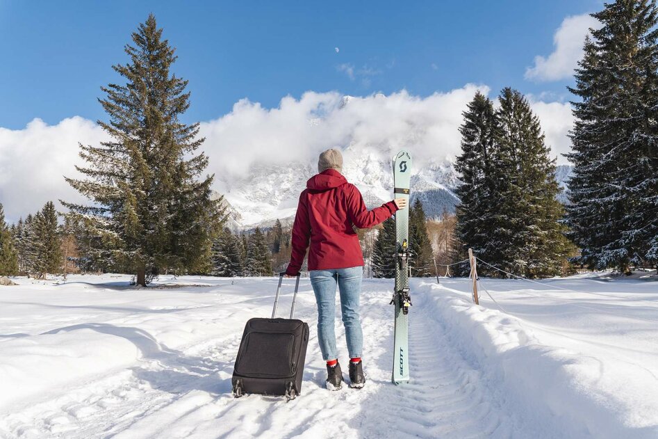 Junge Frau mit einem Koffer und Ski neben sich läuft in Richtung der Zugspitze welche im Hintergrund Sichtbar ist. Die Landschaft ist verschneit. 