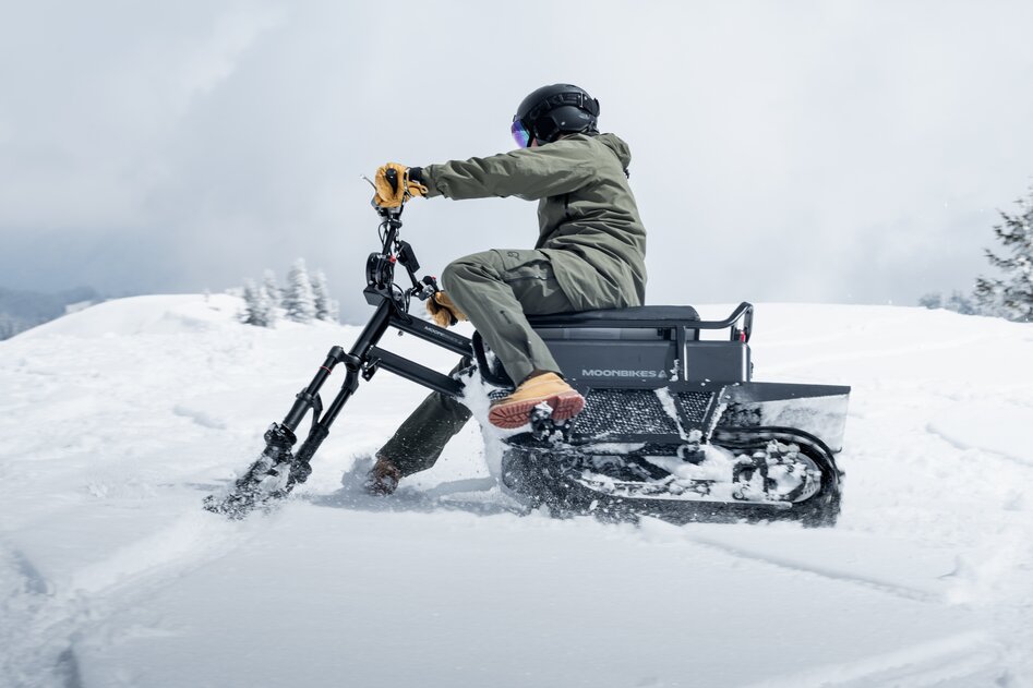 Ein Moonbikefahrer in oliv-grünem Skianzug mit braunen Handschuhen und Schuhen driftet über einen verschneiten Hang mit kleinen Tannenbäumen. 